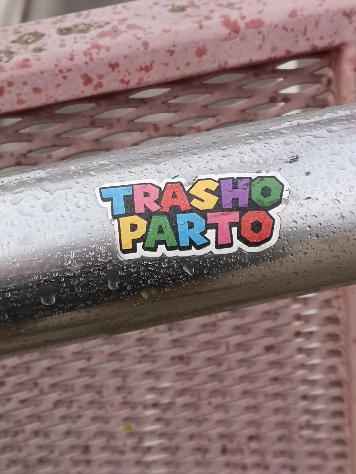 A vibrant, die-cut sticker featuring the words 'TRASHO PARTO' arranged in two lines. The top line reads 'TRASHO' and the bottom 'PARTO'. Each letter is a distinct, brightly colored block (blue, pink, yellow, purple, green, red) with a thick white outline and a thinner black border, strongly reminiscent of the Super Mario Bros. logo font. The sticker is adhered to a wet, metallic, cylindrical surface, likely a railing or pipe, with numerous water droplets visible on both the sticker and the surrounding metal.
