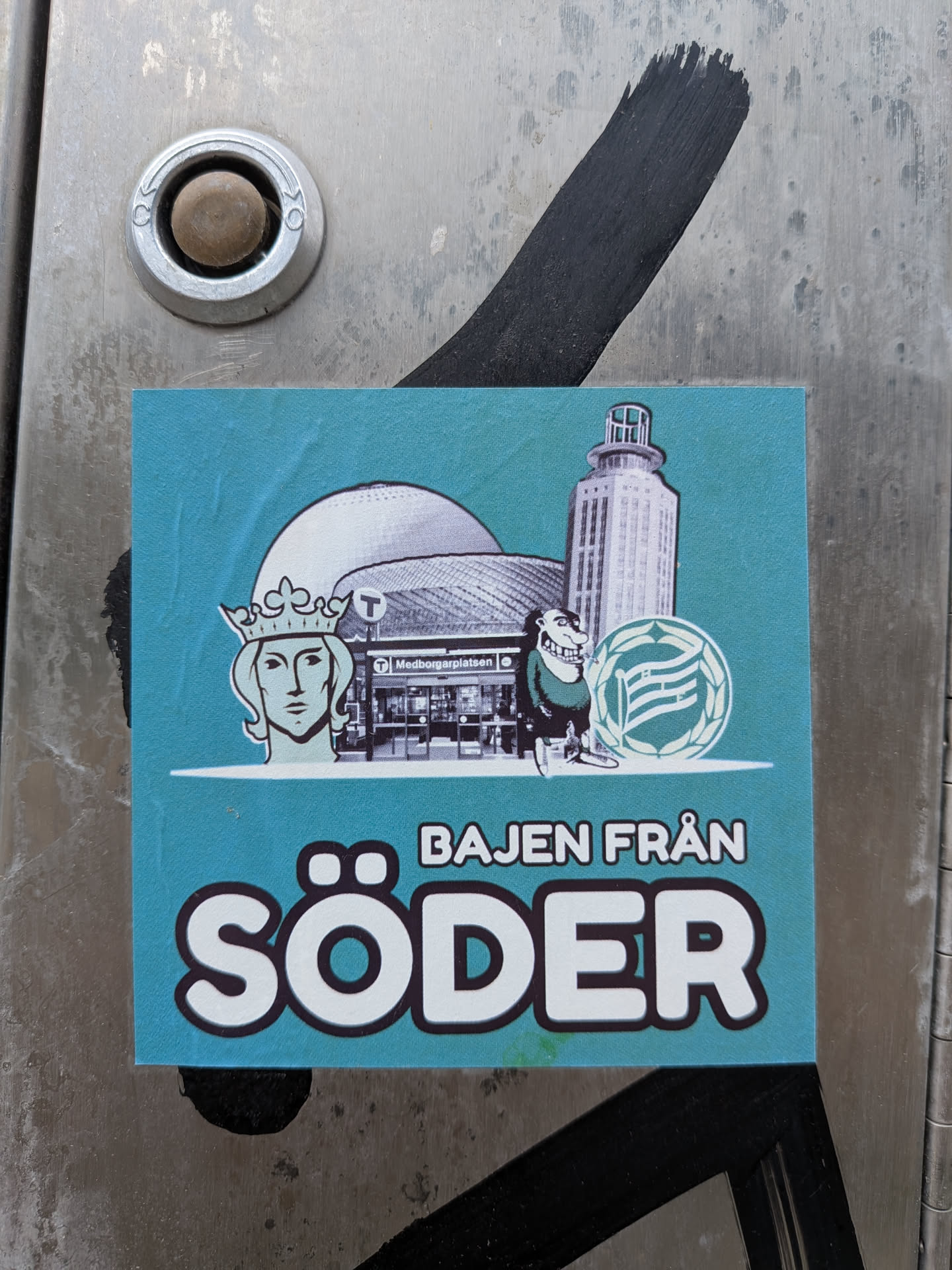 A square teal sticker featuring the text 'BAJEN FRÅN SÖDER' in bold white letters with black outlines. Above the text, a collage of Stockholm and Södermalm landmarks is depicted, including the Globen arena dome, the Medborgarplatsen metro station entrance, the Söder Torn tower, the Stockholm coat of arms (Saint Erik), a cartoon character, and the Hammarby IF club crest.