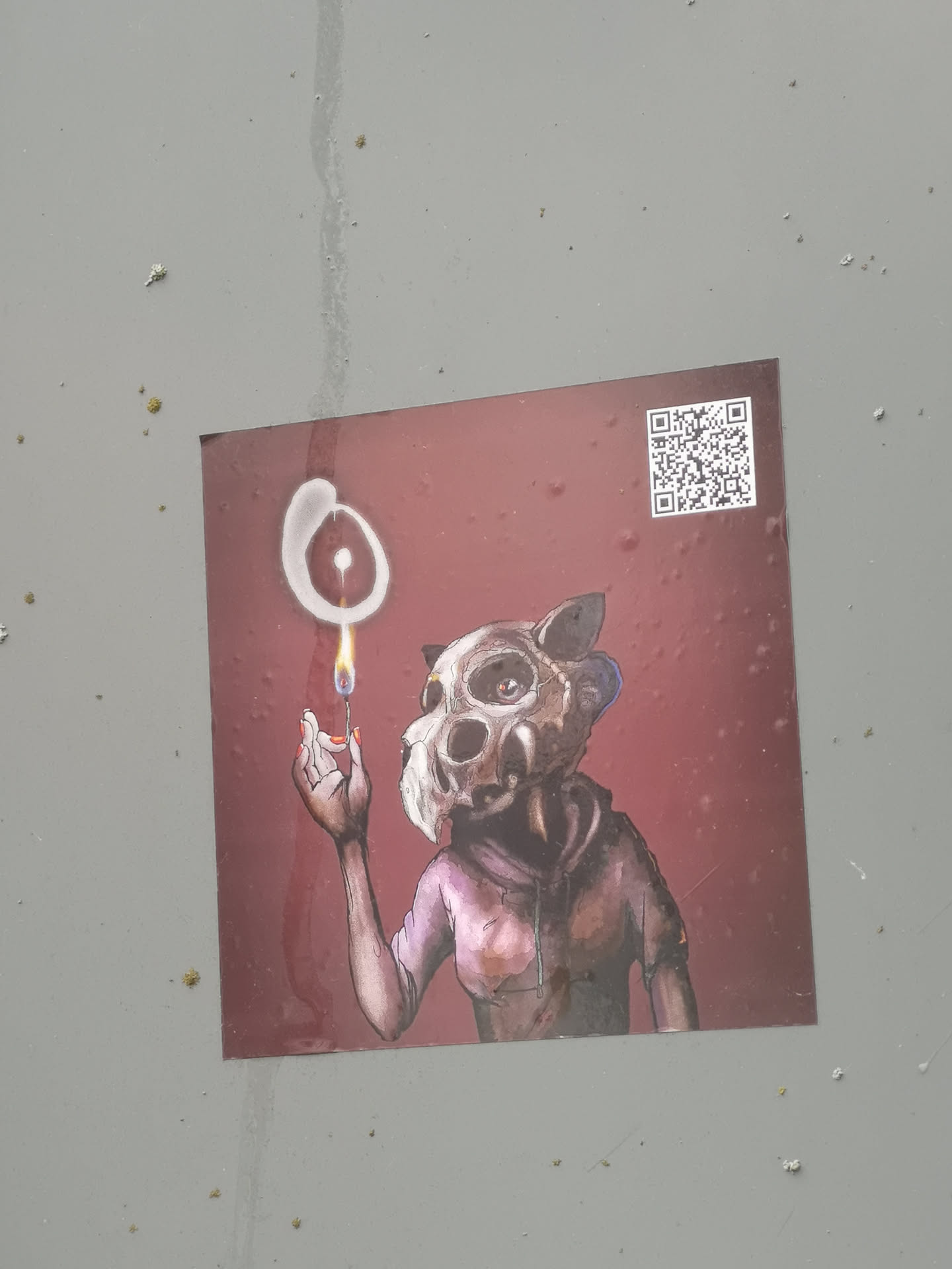 The sticker features a central, stylized figure wearing an animal-like skull mask, possibly resembling a cat or fox, with prominent eye sockets and small ears. The figure is depicted from the chest up, wearing what appears to be a dark, hooded garment in shades of brown and purple. Its right hand, with visible orange-red painted fingernails, holds a lit matchstick. Above the match's flame, a glowing, abstract white symbol, resembling an incomplete circle or an 'O' with a central dot, hovers. The background is a solid, dark reddish-brown color, with subtle textures and imperfections. A QR code is present in the top right corner.