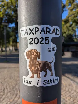 Street sticker The sticker features a stylized illustration of a brown dachshund with a white chest and paws, standing on a white background. Above the dog, the text 'TAXPARD 2025' is prominently displayed in bold black letters. To the right of the text, there are two small black paw prints. Below the dog, the text 'Tax i Sthlm' is written in black letters. The overall shape of the sticker is irregular, with a white border.