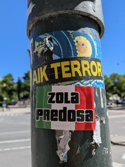 Street sticker The central sticker is rectangular, displaying the vertical stripes of the Italian flag: green on the left, white in the middle, and red on the right. Overlaid on the white and red stripes, and slightly extending into the green, is the text "ZOLA PREDOSA" presented in two lines, with "ZOLA" above "PREDOSA". The text is black, bold, and blocky, with a thin white outline. The sticker is affixed to a dark, weathered metal pole and shows signs of wear, including minor scuffs and slight peeling at its bottom edge.