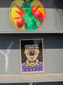 Street sticker A square sticker with a cyclops-like character. The character has one large eye, and is wearing a purple shirt. The background is black with small dots. The text at the bottom of the sticker reads "DOTSY".