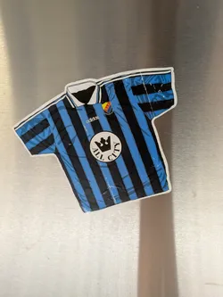 Street sticker The sticker is shaped like a football jersey, predominantly featuring vertical black and blue stripes. It has a white collar with black trim and white cuffs. On the upper right chest, there's a club crest with yellow, red, and blue colors, displaying 'D.I.F.' and 'STOCKHOLMS STÖRSTA'. Below the collar on the left, the 'adidas' logo is visible in white. Centered on the jersey is a large white circular patch with a black crown icon in the middle, surrounded by the text 'ALL CITY' in black capital letters. The sticker has a distinct white outline and shows some minor creases and reflections, indicating its physical nature.