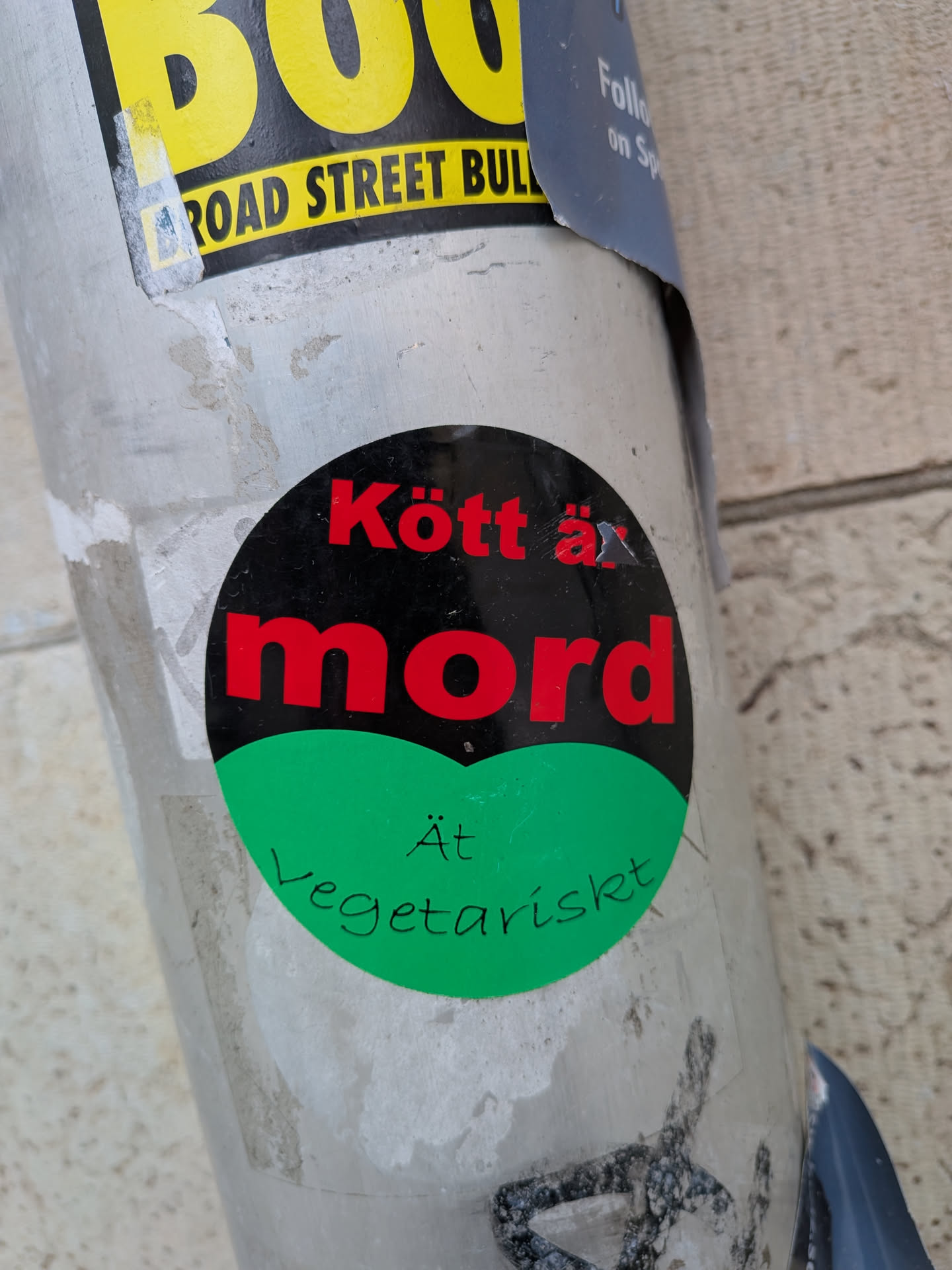 A circular sticker prominently displayed on a grey pole. The top two-thirds of the sticker feature a black background with bold red text that reads 'Kött är mord' (Meat is murder). The bottom third transitions to a bright green background with smaller, handwritten-style black text that says 'Ät Vegetariskt' (Eat Vegetarian). The sticker has a slightly worn appearance, with minor scratches and a small tear near the 'ä' in 'är'. It is partially layered over another yellow sticker visible at the top.