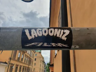 Street sticker A black and white sticker with the text \"LAGOONIZ\" in a stylized font, followed by \"FILM\" in a smaller font. The sticker is applied to a metal pipe outdoors.