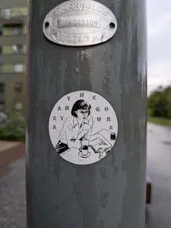 Street sticker The sticker is circular and features a black and white illustration of a woman sitting at a desk, writing or reading. She has short, dark hair and is wearing a collared shirt. In front of her is an open book, a pen, a cup of coffee on a saucer, and another glass. The text 'THE GORGEOUS ANTS' is arranged in a circle around the illustration. The artist's signature 'Kayla Dameltson' is visible in the lower left corner of the illustration. The sticker is slightly wet, with water droplets visible on its surface.