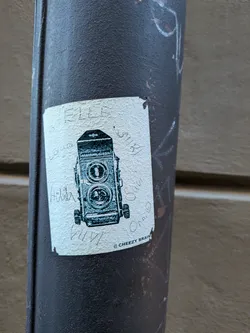 Street sticker A rectangular, off-white sticker featuring a detailed black and white illustration of a Mamiya C330 Professional twin-lens reflex camera. The camera is depicted front-on, showcasing its two prominent lenses, various dials, and the brand name. The sticker has a slightly textured surface and shows signs of wear, with some handwritten names or words like 'ELLE', 'Laura', 'Helia', 'VIVI', 'SIRI', 'Sana', and 'Omni' scribbled around the camera illustration. Below the camera, the text '@ CHEEZY BRAIN' is printed. The sticker is adhered to a dark, heavily textured metal pole.