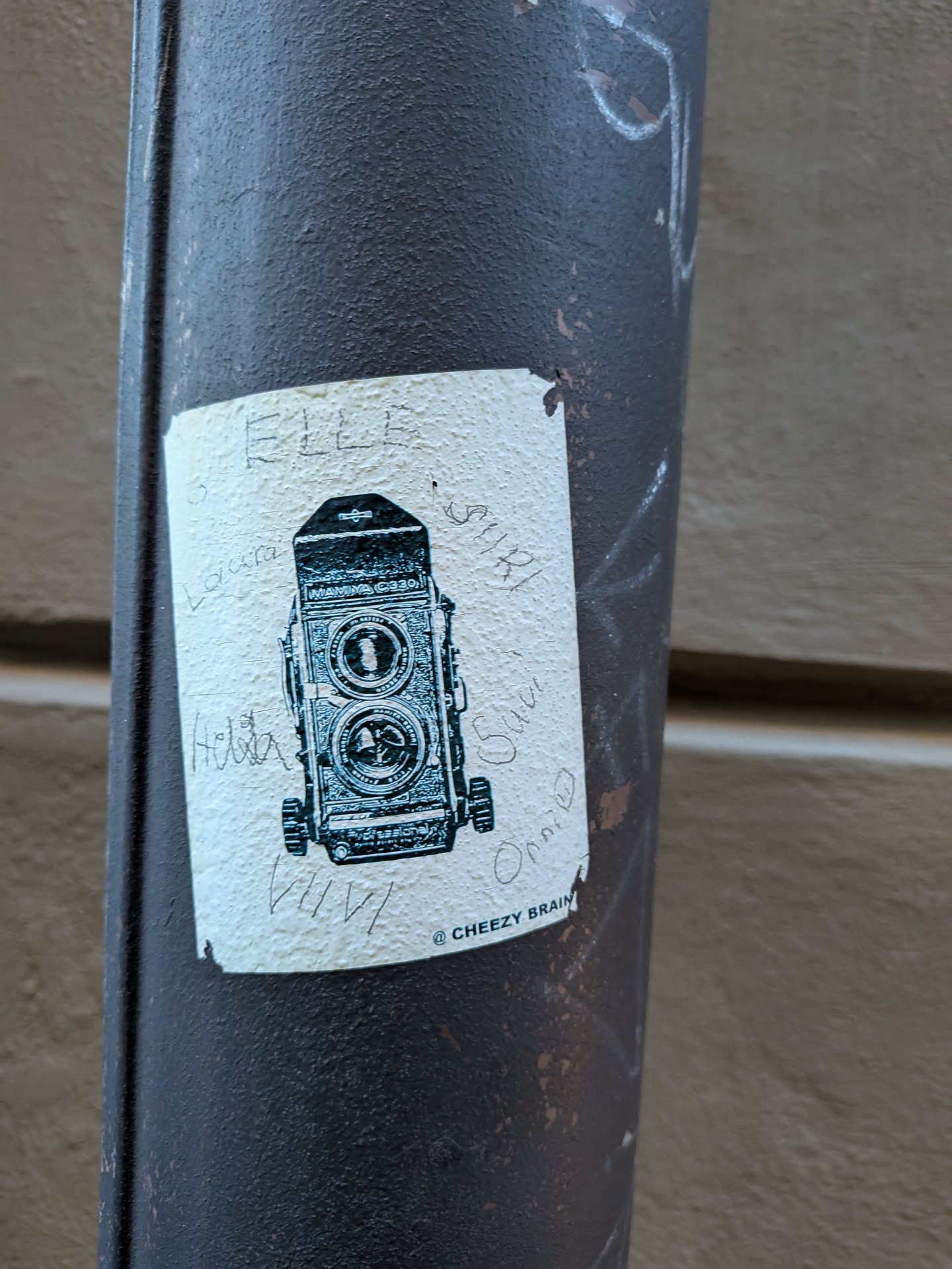 A rectangular, off-white sticker featuring a detailed black and white illustration of a Mamiya C330 Professional twin-lens reflex camera. The camera is depicted front-on, showcasing its two prominent lenses, various dials, and the brand name. The sticker has a slightly textured surface and shows signs of wear, with some handwritten names or words like 'ELLE', 'Laura', 'Helia', 'VIVI', 'SIRI', 'Sana', and 'Omni' scribbled around the camera illustration. Below the camera, the text '@ CHEEZY BRAIN' is printed. The sticker is adhered to a dark, heavily textured metal pole.