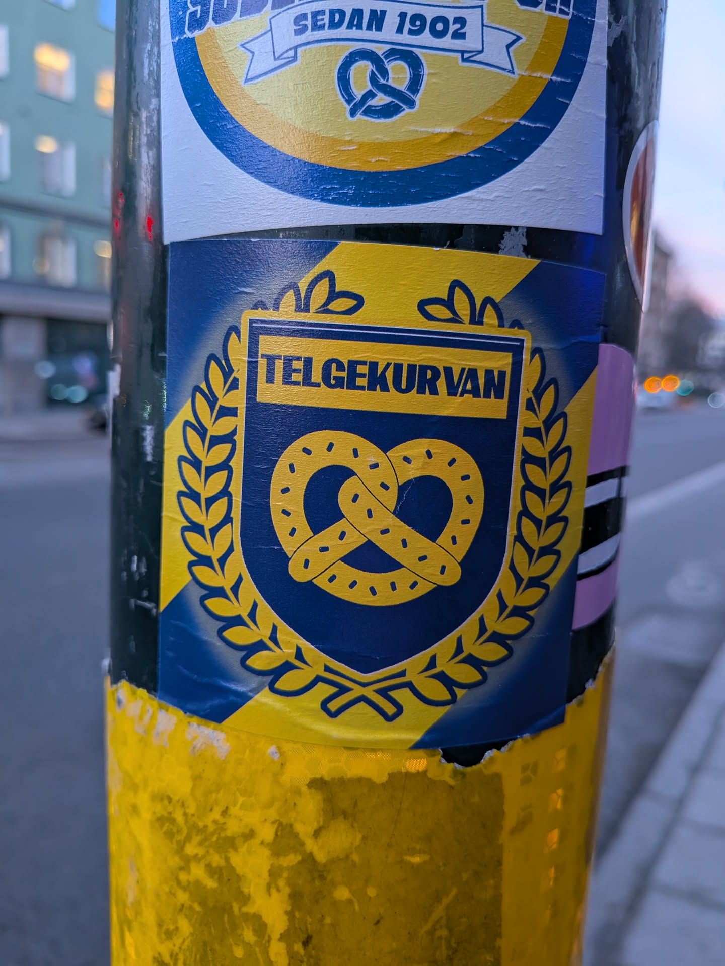 Street sticker The sticker features a prominent blue shield with a yellow pretzel at its center. The pretzel is outlined in dark blue and dotted with small yellow circles. The shield is surrounded by a laurel wreath in yellow. Above the pretzel, within the shield, is a yellow banner with the text 'TELGEKURVAN' in dark blue capital letters. The background of the sticker is a gradient of yellow and blue, with diagonal stripes. The edges of the sticker are slightly torn and peeling, indicating it has been applied to a surface.