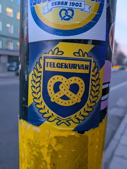 Street sticker The sticker features a prominent blue shield with a yellow pretzel at its center. The pretzel is outlined in dark blue and dotted with small yellow circles. The shield is surrounded by a laurel wreath in yellow. Above the pretzel, within the shield, is a yellow banner with the text 'TELGEKURVAN' in dark blue capital letters. The background of the sticker is a gradient of yellow and blue, with diagonal stripes. The edges of the sticker are slightly torn and peeling, indicating it has been applied to a surface.