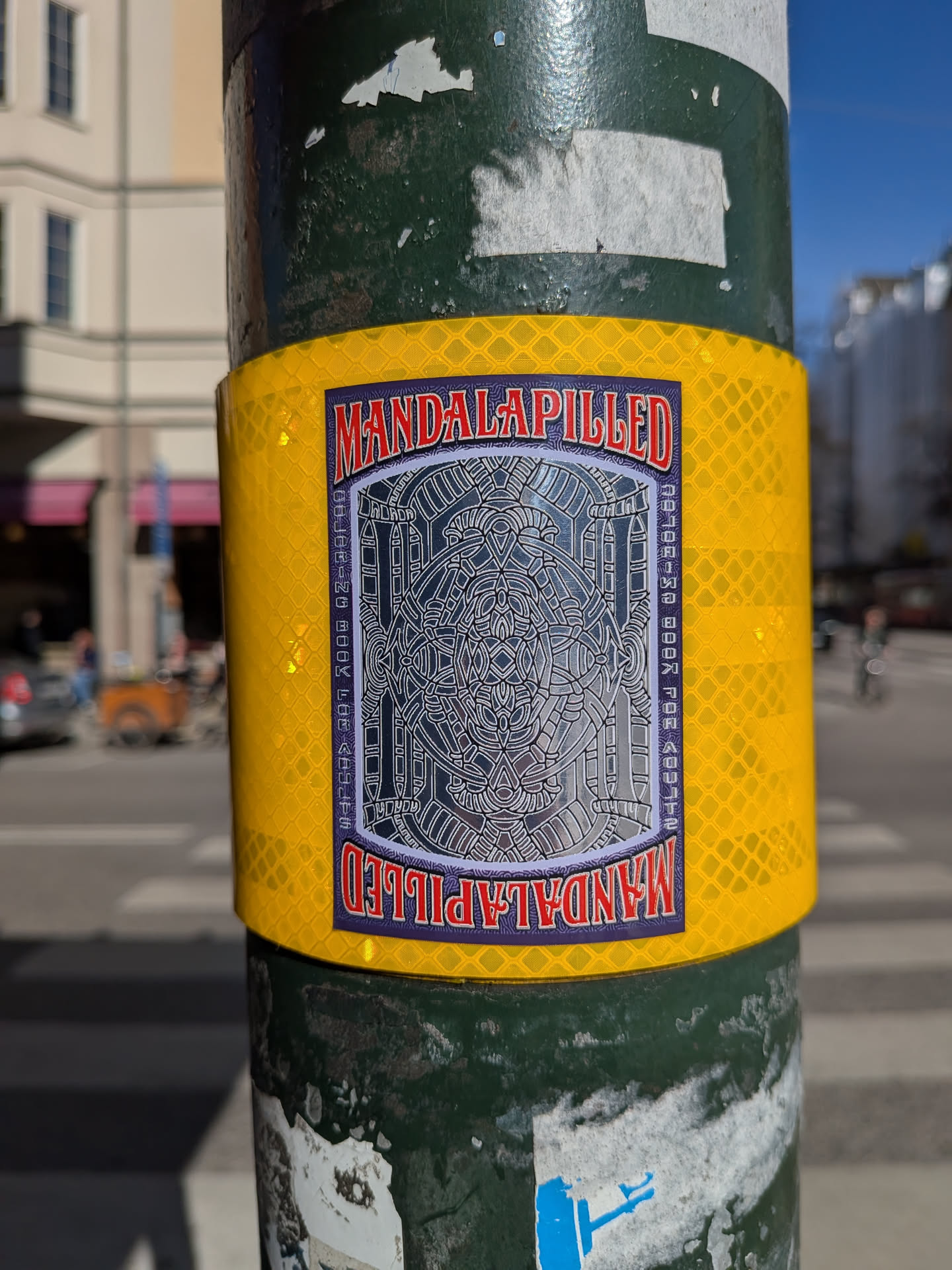 The sticker is rectangular with slightly rounded corners, featuring a dark purple border. It is prominently displayed on a bright yellow, reflective, diamond-patterned surface wrapped around a pole. At the top and bottom, a red banner with a white outline contains the word "MANDALAPILLED" in a bold, stylized font; the text at the bottom is inverted. The central area of the sticker is dominated by a highly intricate, symmetrical geometric pattern or mandala rendered in shades of silver and grey, giving it a metallic or embossed appearance. Along the vertical sides of this central design, in smaller white text against the purple border, are the words "COLORING BOOK FOR ADULTS".