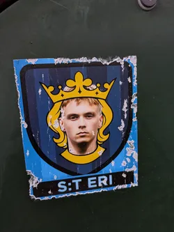 Street sticker The sticker features a stylized shield with vertical dark blue stripes. At the top center is a prominent golden crown with intricate details. Within the shield, a young man with blond hair and a serious expression is depicted, his head framed by the crown and a yellow outline. Below the portrait, a black band contains the text 'S:T ERI' in light blue. The edges of the sticker are worn and torn, with visible peeling and white paper backing.