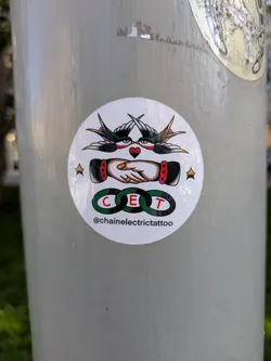 The sticker is circular with a white background. At the top, two stylized swallows in black and red with yellow accents are depicted in mid-flight, facing each other, with their wings extended. Between them, a red heart with black outlines is positioned. Below the swallows, two hands are shaking, rendered in a traditional tattoo style with detailed shading and a red cuff on one wrist. Encircling the center of the handshake are three interlocking rings, each with a green outline and a letter inside: 'C', 'E', and 'T'. Below the rings, the text '@chainelectrictattoo' is visible in black lettering. The overall aesthetic is reminiscent of traditional tattoo flash art.