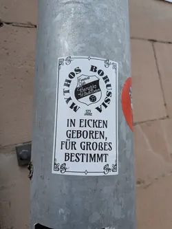 Street sticker The sticker is a rectangular design with an ornate, decorative black border on a white background. At the top, the words "MYTHOS BORUSSIA" are arched in bold black capital letters. Centered below this is a shield-shaped crest, predominantly black with white text, featuring "Fussball Club" at the top, "Borussia" in the middle, and "M.Gladb. Eicken" below. A small football icon is positioned beneath the crest, followed by the text "125 JAHRE". The lower section of the sticker displays a three-line German motto in bold black capital letters: "IN EICKEN GEBOREN,", "FÜR GROSSES", "BESTIMMT". The sticker shows signs of being outdoors, with visible water droplets on its surface, and is affixed to a textured grey metallic pole.