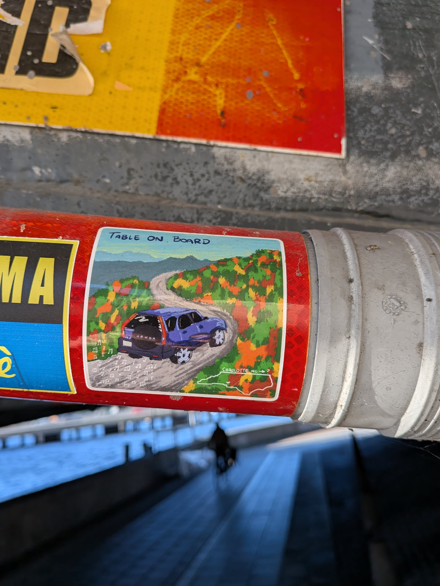 The sticker features a vibrant illustration of a blue Honda CR-V driving on a winding road through a colorful landscape of autumn trees and mountains. Musical notes are scattered around the car, suggesting music is playing. The bottom of the sticker shows a map outline of North Carolina with 'CHARLOTTE NC' labeled. The sticker has a red, textured background with some text and graphics on the left side that are partially obscured.