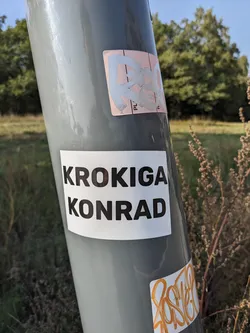 A minimalist sticker with the names Krokiga Konrad printed in bold black letters on a white background. The sticker is affixed to a gray pole, surrounded by other smaller stickers and graffiti.
