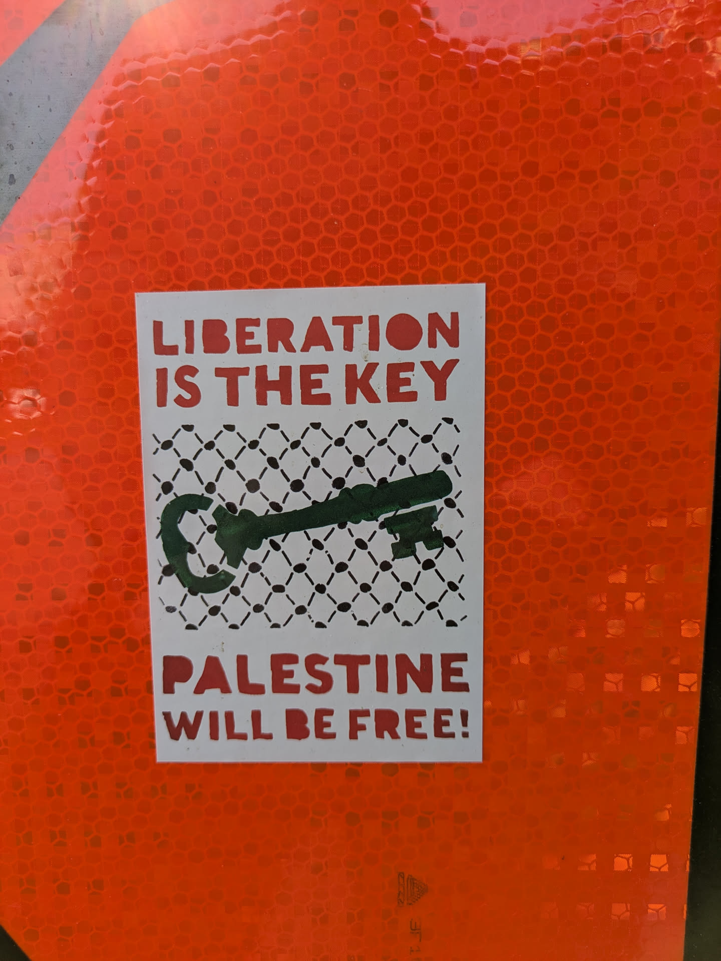 This sticker features a green key against a black and white keffiyeh pattern. The text reads 'LIBERATION IS THE KEY' and 'PALESTINE WILL BE FREE!' in bold red letters. The sticker is adhered to an orange, reflective surface.