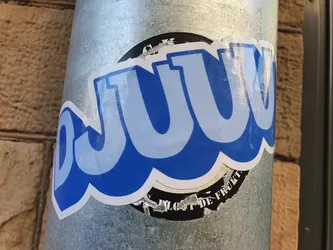 Street sticker A blue and white sticker with the word "DJUUW" in a stylized font. The sticker is applied to a metallic cylindrical surface, likely a pipe or drainpipe.  There is a smaller, partially visible, black and white sticker underneath.