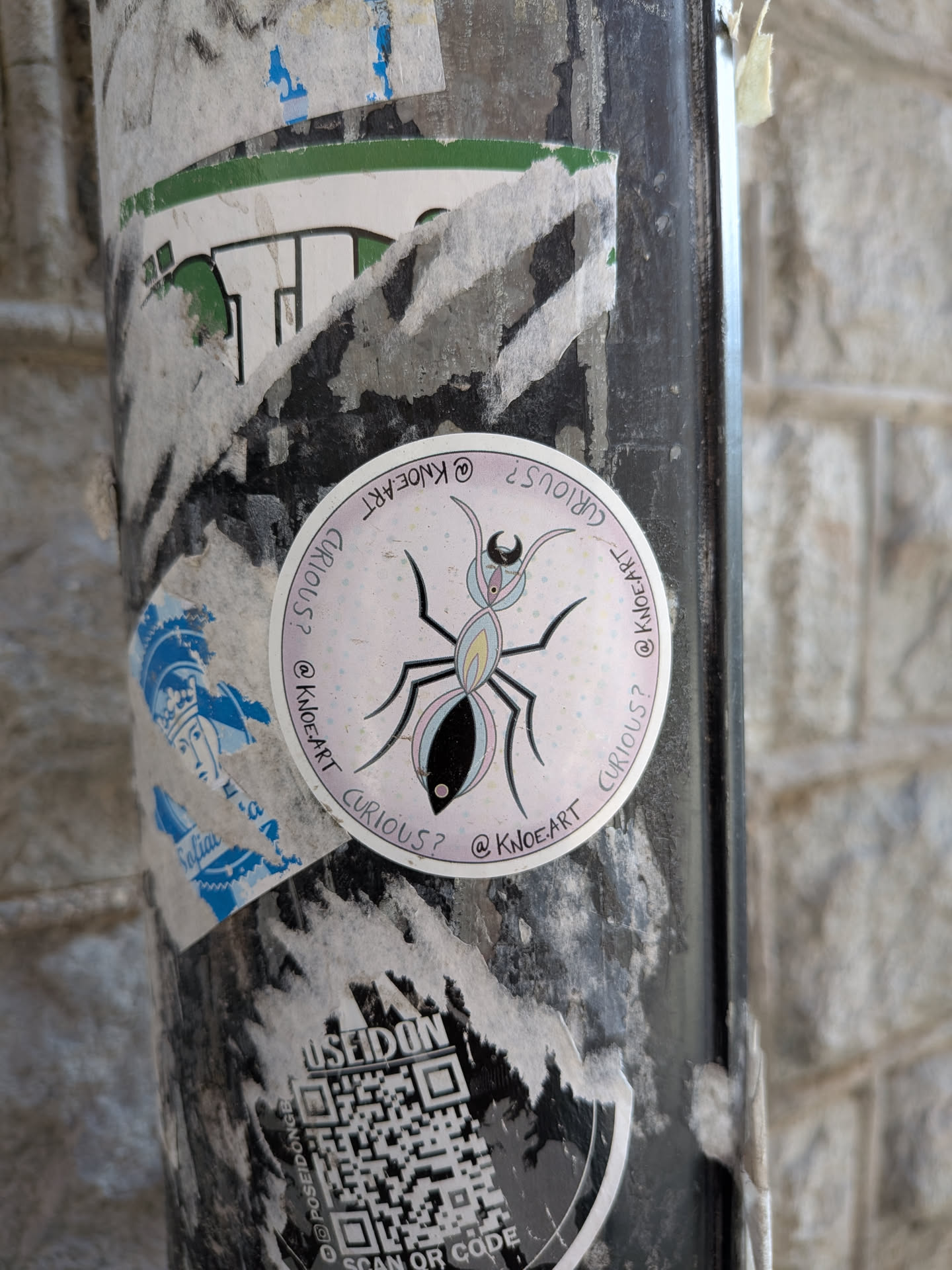 A circular sticker featuring a stylized ant illustration against a light pink background speckled with small, colorful dots (yellow, blue, pink). The ant has a black head with two small antennae, a light blue thorax with six black legs, and an abdomen divided into a light yellow front section and a black rear section. The entire ant is outlined in black. Around the perimeter of the sticker, the words "CURIOUS?" and "@KNOE.ART" are repeated in black text, alternating.