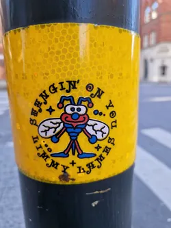 Street sticker A yellow and black circular sticker features a cartoon bee with the text 'Hangin' on my nose' surrounding it. The bee is blue, red, and white, with simple, cartoonish features. The style is reminiscent of vintage or retro designs. The sticker is applied to a dark grey pole, possibly in an urban setting.