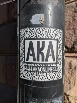 A rectangular sticker with slightly rounded corners, featuring a high-contrast black and white design. The outer border of the sticker has a white background with an irregular, organic black pattern, reminiscent of camouflage or abstract animal print. Centered within this pattern is a solid white rectangular box with a thick black outline. Inside this box, the large, bold, sans-serif letters 'AKA' are displayed in black, with small black dots filling the negative space of the 'A's. Below the 'AKA' text, in a smaller black sans-serif font, is the website 'WWW.AKAONLINE.SE'. The sticker is applied to a dark, textured, and somewhat weathered pole or pipe, showing minor wear and scratches.