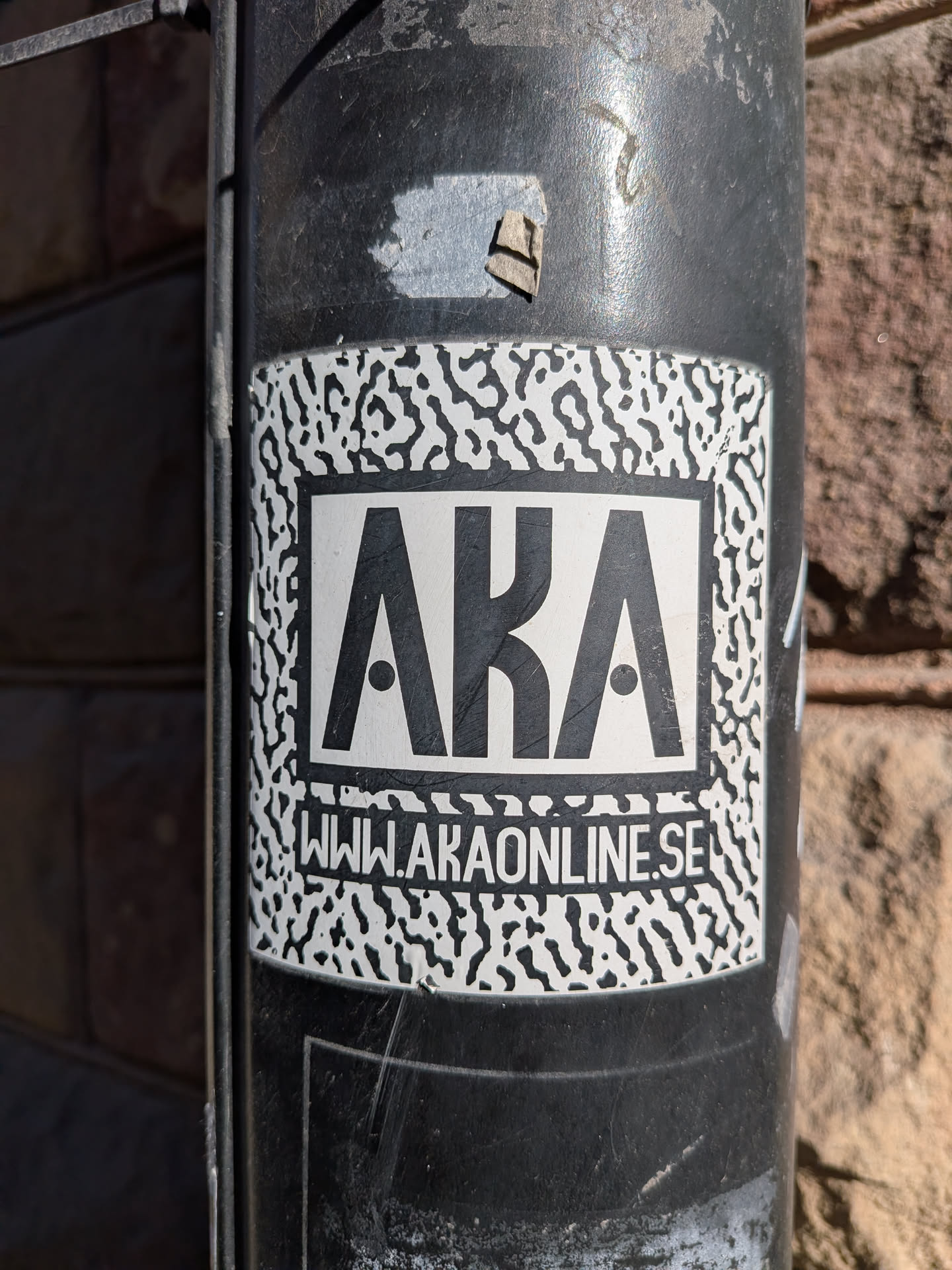 A rectangular sticker with slightly rounded corners, featuring a high-contrast black and white design. The outer border of the sticker has a white background with an irregular, organic black pattern, reminiscent of camouflage or abstract animal print. Centered within this pattern is a solid white rectangular box with a thick black outline. Inside this box, the large, bold, sans-serif letters 'AKA' are displayed in black, with small black dots filling the negative space of the 'A's. Below the 'AKA' text, in a smaller black sans-serif font, is the website 'WWW.AKAONLINE.SE'. The sticker is applied to a dark, textured, and somewhat weathered pole or pipe, showing minor wear and scratches.