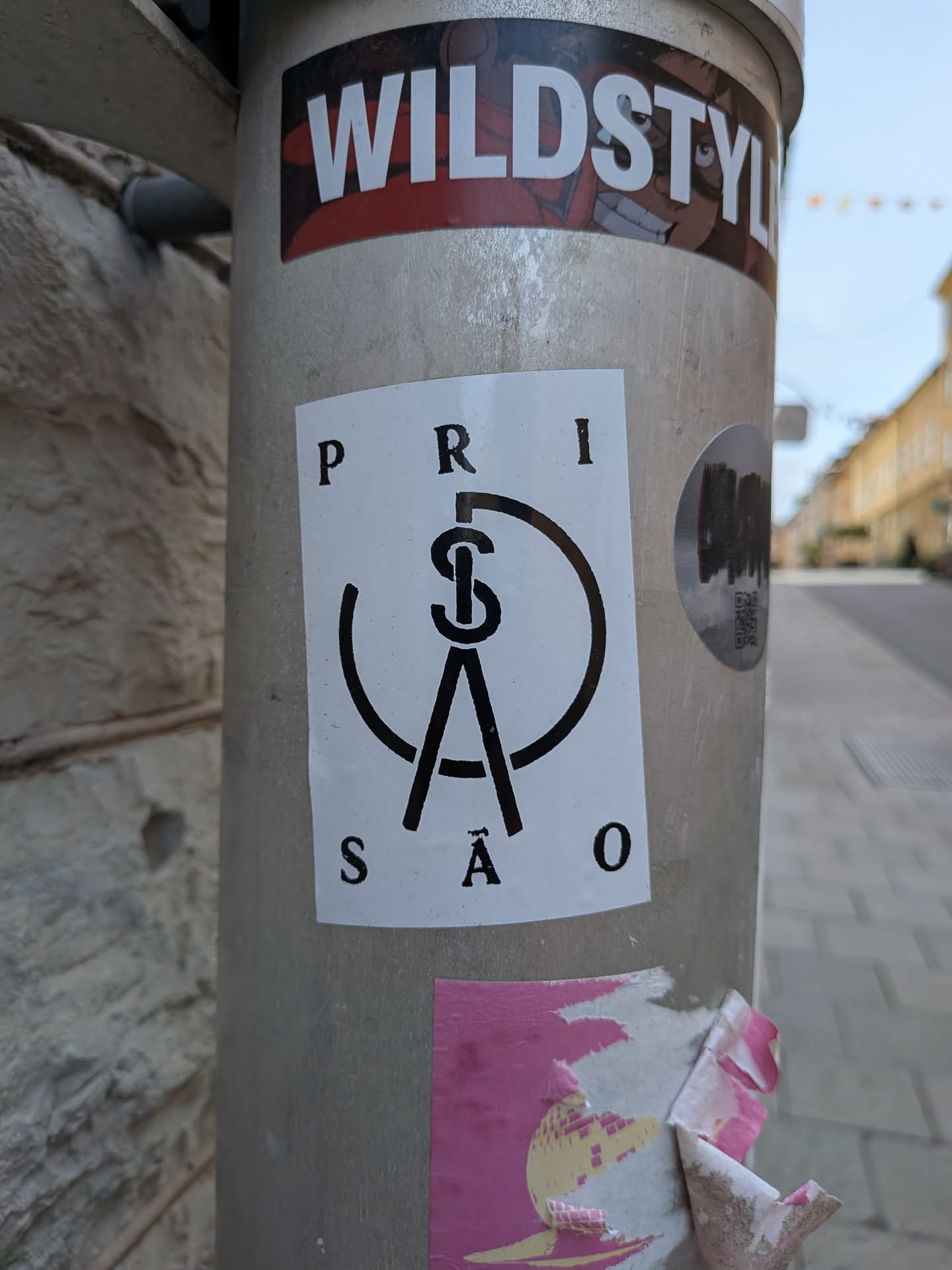 A white rectangular sticker featuring the black text 'PRISÃO' split top and bottom, surrounding a central black monogram logo of intertwined letters within a broken circle.