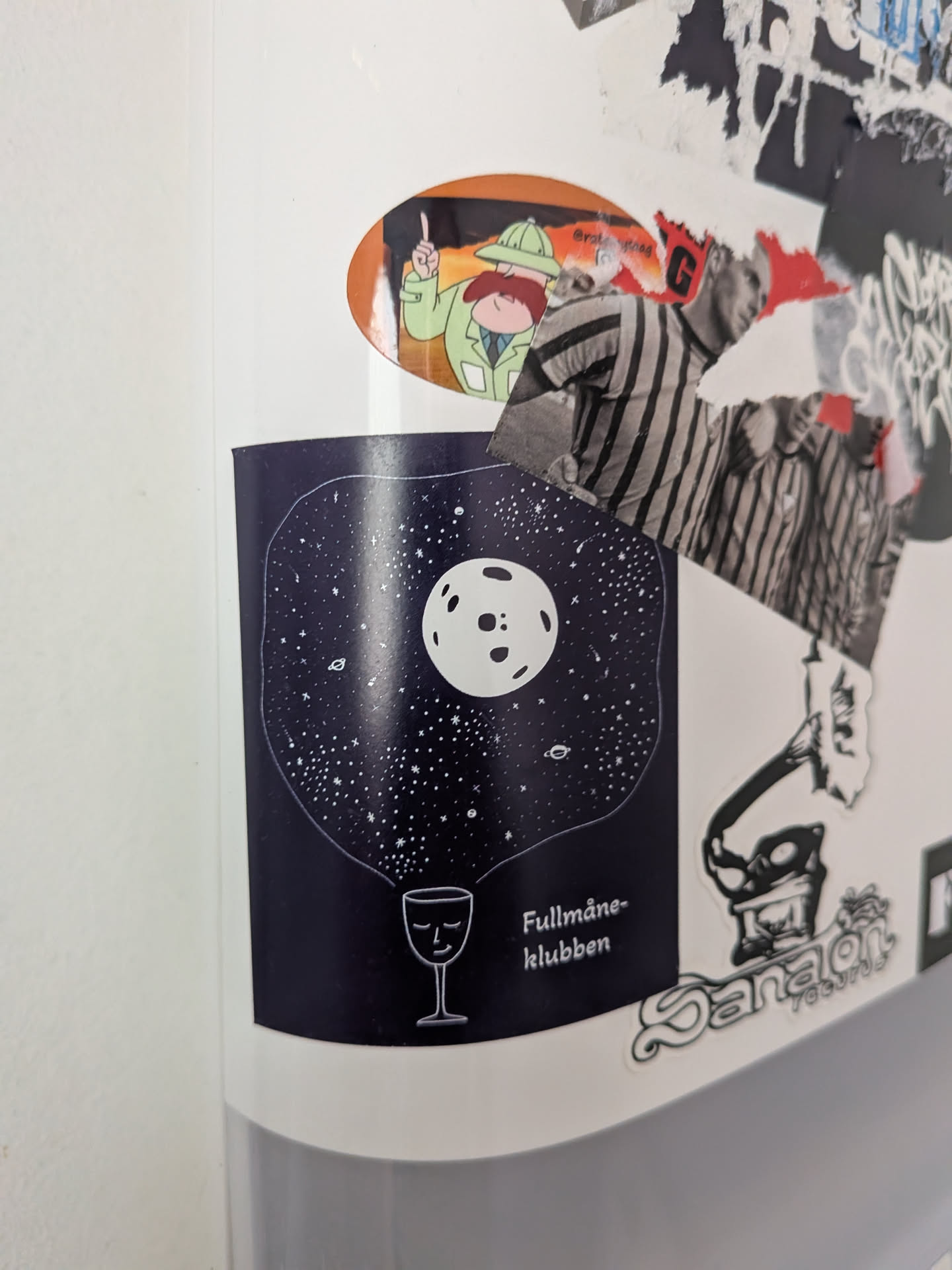 A dark blue rectangular sticker with a rounded top edge, featuring a minimalist illustration of a night sky. A prominent white full moon with visible craters is centered in the upper half, surrounded by numerous small white stars and a few tiny planetary bodies. Below this celestial scene, a simple white outline of a wine glass is depicted, with a serene, closed-eye face. To the right of the wine glass, the white text 'Fullmåne-klubben' is present. The overall aesthetic is dreamy and cosmic.