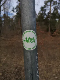 Street sticker A circular white sticker featuring green silhouettes of diverse figures walking together. The group includes a dog, several adults, one of whom is in a wheelchair, and a child. The figures are stylized and appear to be in motion. Encircling the central image is green text in Swedish. The sticker is affixed to a weathered, grey metal pole, showing some minor surface imperfections and wrinkles.