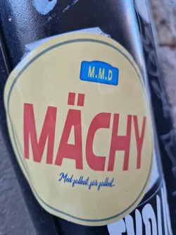 Round sticker with the text "Mächy" in bold red letters on a pale yellow background.  A smaller blue rectangle at the top says "M.M.D."  Under the main text is a smaller line in a cursive blue script that reads "Med folket, för folket".