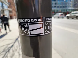 A black and white sticker featuring the logo for Back2Back Records, depicting two turntables.  The text 'BACK2BACK RECORDS' and 'STHLM' are visible on the sticker. The sticker is affixed to a dark grey pole on a city street. 