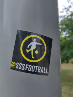 A square sticker featuring a simple graphic of a soccer player kicking a ball, enclosed in a yellow circle on a black background. The text '@SSSSFOOTBALL' is visible at the bottom. The sticker is affixed to a gray pole outdoors.