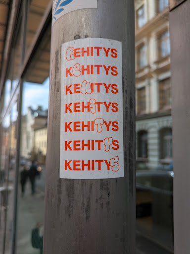The sticker features the word "KEHITYS" printed in orange multiple times on a white background, affixed to a metal pole.
