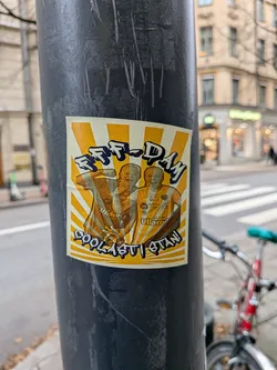 Street sticker The sticker features two individuals, likely football fans, depicted in a stylized manner. They are wearing jerseys with visible team logos and text. The background is a radiating pattern of yellow and white stripes, creating a sense of energy and excitement. The overall color palette is dominated by yellow, orange, and brown tones, with black outlines and text. The individuals appear to be cheering or celebrating.