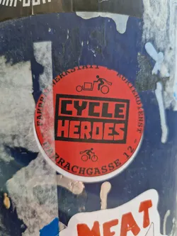 Street sticker Red and black circular sticker with the text "CYCLE HEROES" in bold letters. It also contains a small illustration of a person riding a bicycle with a cargo trailer. The text around the circle seems to indicate a location or address: "FAPPER MERKERSTAATT KURIERDIENST - ULRACHGASSE 12"