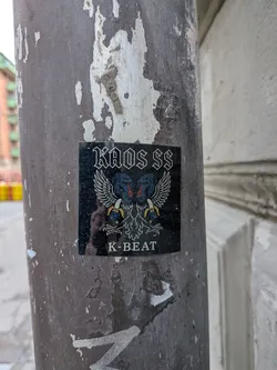 Street sticker A square, dark-themed sticker featuring a central graphic of two mirrored, winged baboon-like creatures. The baboons have blue faces with red accents, white feathered wings spread wide, and each holds a yellow banana in its clawed hand. Above the graphic, the text "KAOS SS" is written in a stylized, blocky, somewhat gothic font, with the 'SS' resembling lightning bolts. Below the graphic, the text "K-BEAT" is displayed in a simpler, sans-serif white font. The sticker has a slightly glossy finish, reflecting some light and a faint silhouette of the photographer.