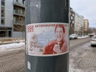 Street sticker The sticker is designed to resemble an old banknote, predominantly in off-white, red-orange, and blue-grey tones. It features a prominent portrait of a smiling man, possibly a caricature, rendered in a red-orange, engraved style. He wears a collared shirt with the number '1891' on it and appears to be holding something near his chest. Behind him, in a blue-grey, finely detailed engraving style, is an illustration of a large, traditional building, possibly a farm or manor, with smaller figures and horses in the foreground. Large red numbers '1891' are visible on the left side, with the text 'SVERIGES BÄSTA LAG' beneath them. At the top, 'ARTONHUNDRA NITTIOETT' is written in red. Smaller '1891' and a numerical sequence '138501891' are also present at the bottom left. The sticker has a slightly worn appearance, adhering to a dark grey pole.