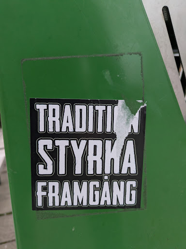 The sticker is black and white and has the words "TRADITION STYRKA FRAMGANG" written in capital letters. It is on a green surface, possibly a trash can or similar object.