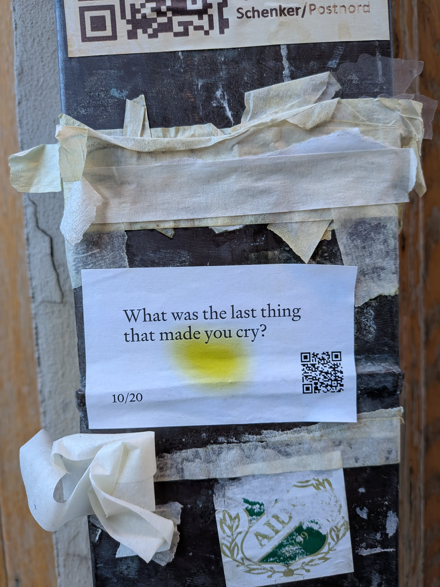 The sticker is a rectangular piece of white paper, affixed to a dark, weathered wooden post with several strips of light-colored masking tape. Centered on the paper is the question 'What was the last thing that made you cry?' printed in black serif font. Below this text, slightly to the left, is the number '10/20'. A prominent, irregular yellow stain or splash is visible in the center of the paper, beneath the main text. In the bottom right corner, a black and white QR code is printed.