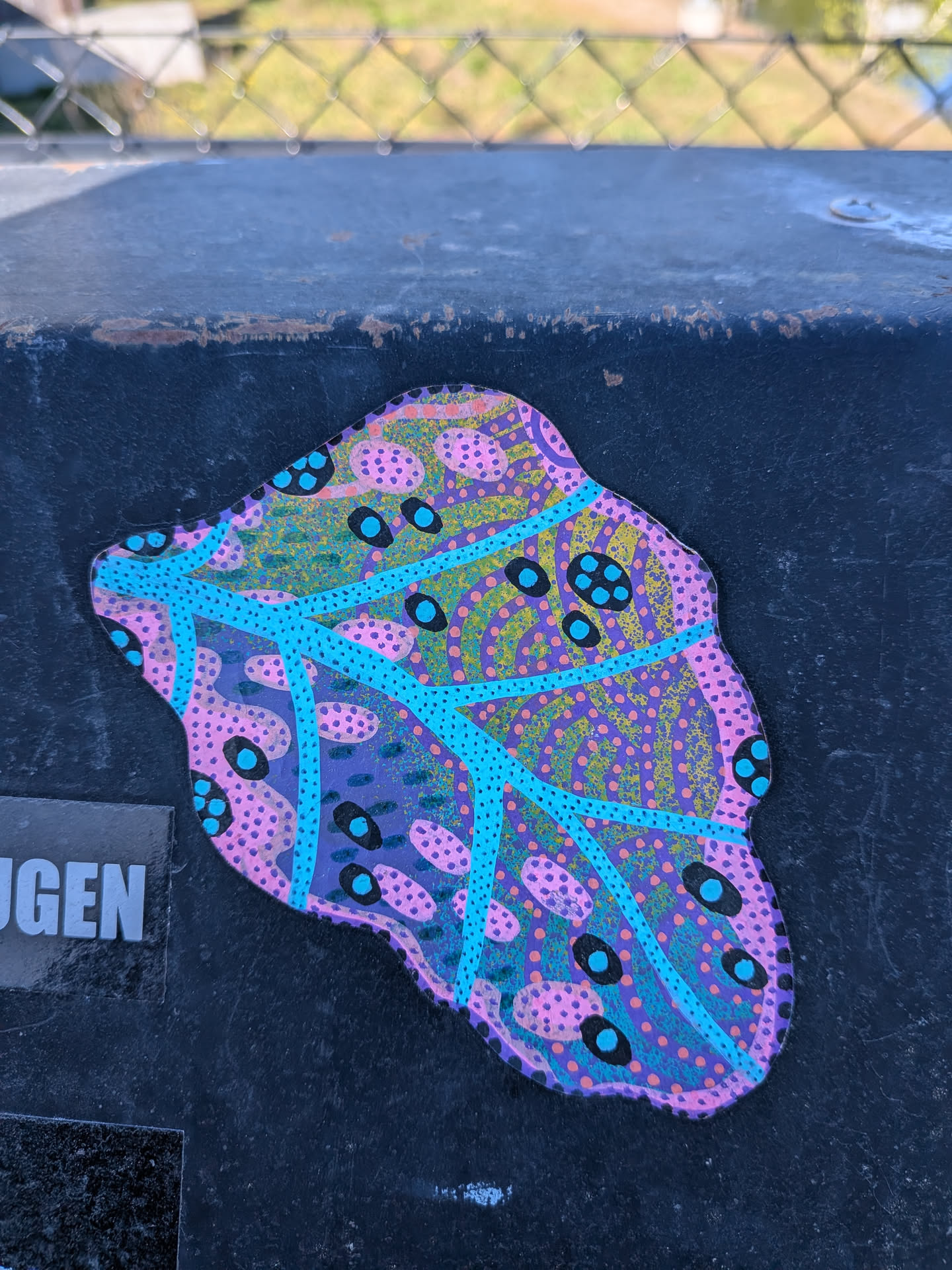The sticker features an irregular, organic, amoeba-like shape, predominantly colored in shades of deep purple, light pink, bright cyan (light blue), and yellow-green. The purple serves as a base, overlaid with intricate patterns. Numerous small pink dots are clustered into oval shapes, while tiny yellow-green dots are scattered throughout. A network of bold, bright cyan lines crisscrosses the surface, resembling veins or a structural framework. Within various sections, larger black oval shapes are present, some containing smaller cyan dots and others containing pink dots. The overall design is a vibrant, layered abstract pattern with a distinct hand-drawn or painted aesthetic, giving it a cellular or microscopic appearance. It is affixed to a dark, textured surface.
