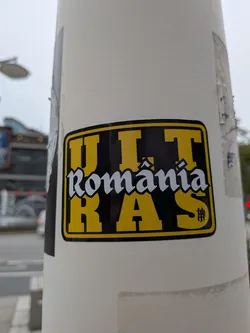 Street sticker A rectangular sticker with rounded corners, featuring a black background and a yellow border. The word 'ULTRAS' is split into 'ULT' at the top and 'RAS' at the bottom in large, bold yellow capital letters. Across the center, the word 'România' is written in a stylized white gothic font. A small black monogram symbol is visible inside the letter 'S'.