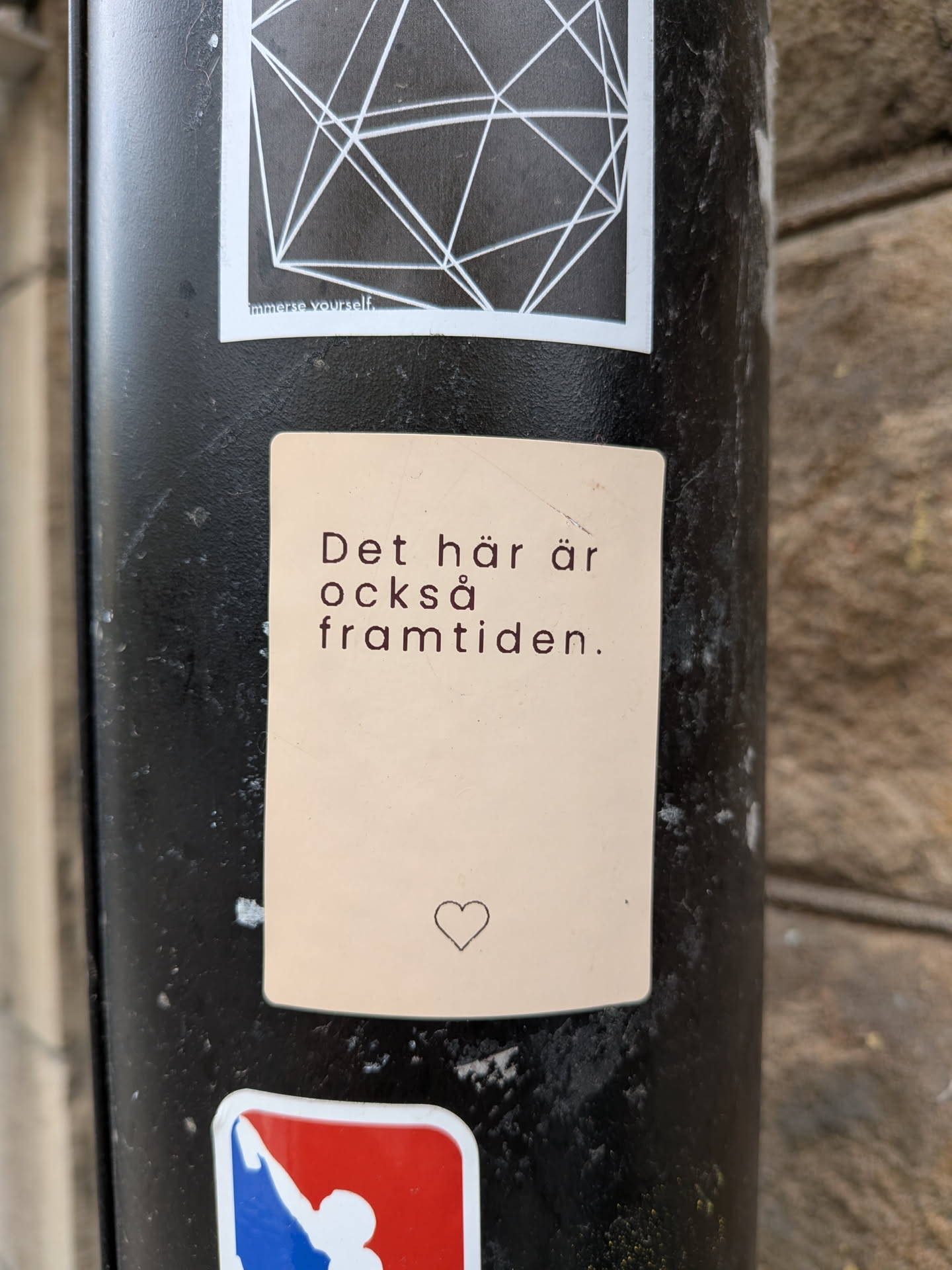 The sticker is a light beige or off-white rectangle with slightly rounded corners. It features dark brown or black text in a clean, sans-serif font, centered on the upper half. Below the text, towards the bottom center, is a small, outlined heart symbol. The sticker has a matte finish and appears to be adhered to a dark, textured pole.