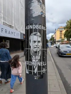 Street sticker The sticker is predominantly black and white, featuring a stylized portrait of a man's face, framed by vertical white bars that resemble prison bars. The man is looking directly at the viewer. Above his head, the name "KINDBERG" is printed in large white capital letters. Below the portrait, the Swedish text "ÄR KRIMINELL" is written, also in white capital letters. The overall impression is stark and impactful, with a hint of wheat-paste style application, suggesting it's a political commentary sticker.