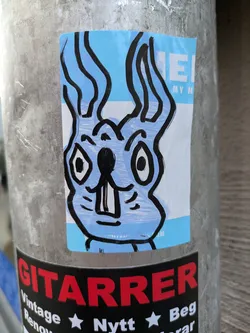 Street sticker This sticker features a light blue rabbit with black outlines. The rabbit has large eyes and a simple, cartoonish expression. The sticker is on a light blue background with some text visible in the upper right corner that reads 'MY N'. The background is mostly blue but has a horizontal white bar under the top blue part.