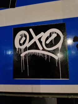 This square sticker features a graphic design in white paint on a black background. The design resembles a face with two large circular eyes and a simple curved line representing the mouth. White paint drips down from the figure, adding a dynamic feel. The sticker has a clean, glossy finish, and the graphic is bold and expressive, creating a striking visual contrast.