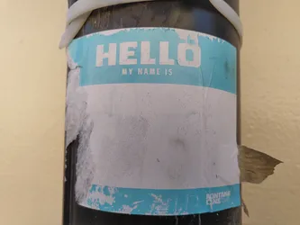 Street sticker A light teal and white name tag sticker is affixed to a dark pole.  The sticker is peeling and worn, with significant damage and dirt. The top portion says "HELLO MY NAME IS" in a sans-serif font. The bottom portion is mostly blank but has a small amount of text visible at the bottom right, possibly a brand or logo.