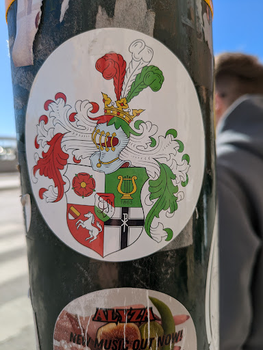 The sticker features a coat of arms with a helmet, feathers, and a crown on top. The shield is divided into sections with different symbols, including a horse, a rose, and a lyre with the year 1863. There is a religious symbol to the right of the lyre. The sticker is round and affixed to a dark green pole.
