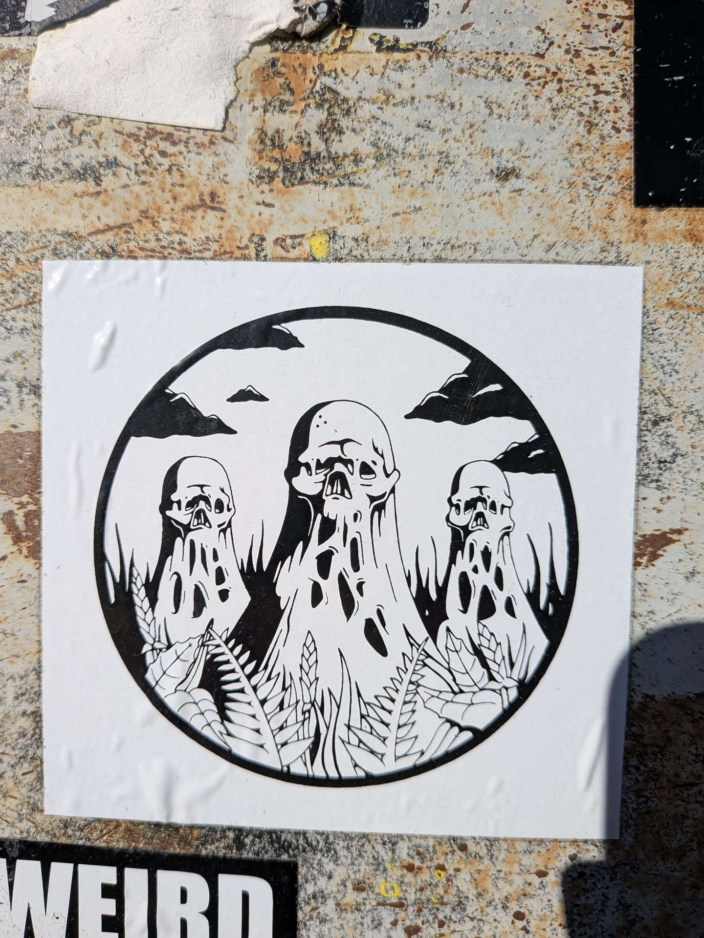 The sticker features a circular, black-bordered illustration on a white rectangular background. Inside the circle, three skull-like figures dominate the scene, rendered in black and white. The central figure is the largest, with two smaller ones on either side. All three appear to be melting or dripping downwards, forming elongated, organic shapes with irregular holes and voids, giving them a ghostly or decaying appearance. The skulls have visible eye sockets and nasal cavities. The base of the figures and the foreground are filled with black silhouettes of various plant life, resembling grass and ferns. In the upper portion of the circle, against the white background, are dark, stylized clouds and small, distant hill-like shapes, all rendered in black. The overall aesthetic is dark, macabre, and surreal, utilizing only black and white tones.