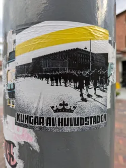 Street sticker The sticker features a striking monochromatic image with a bold diagonal yellow stripe across its top portion, resembling a flag or banner. Below the stripe, a large crowd of people is depicted marching on a wide street. The figures are silhouetted in dark tones against a lighter grey background, with some individuals raising their arms. In the background, dark, imposing buildings with numerous windows form a cityscape. Below the main image, centered, is a stylized black crown with white details. Beneath the crown, there's text in a bold, blocky font with a white outline against a black, speckled background that transitions from the street scene. The overall style is gritty and impactful, suggesting a scene of movement or a rally.