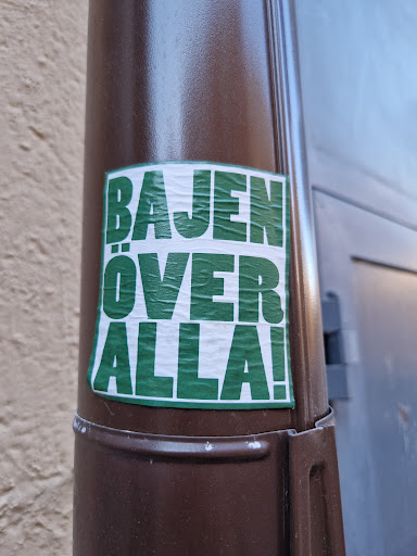 Street sticker A green sticker with white text on a brown pole. The text says "BAJEN ÖVER ALLA!"